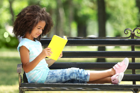 school age girl reading book on park bench